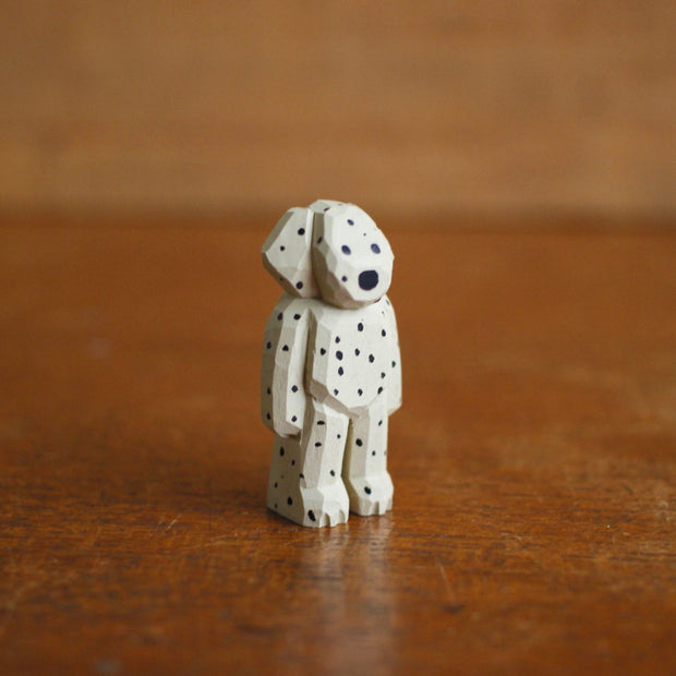 Tiny painted whittled wooden sculpture of a dog with floppy ears, standing on its hind legs like a human. Its white with many black spots, and a small pair of black eyes and a large nose.