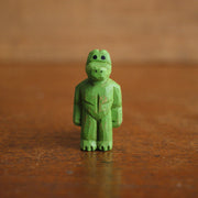 Small painted whittled wooden sculpture of a green gator, standing on 2 legs like a human with a strong body. It has a blank expression and its arms hang at its sides.