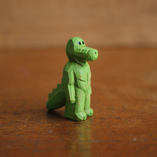 Small painted whittled wooden sculpture of a green gator, standing on 2 legs like a human with a strong body. It has a blank expression and its arms hang at its sides.