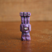 Small whittled wooden sculpture of a purple alien, with 3 eyes extended out from its head. Its body just contains its mouth, smiling with buckteeth, and it stands on 2 legs without arms.