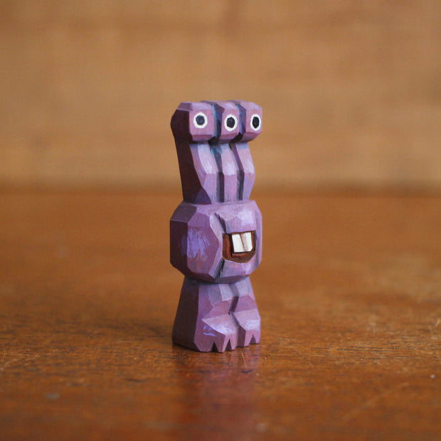 Small whittled wooden sculpture of a purple alien, with 3 eyes extended out from its head. Its body just contains its mouth, smiling with buckteeth, and it stands on 2 legs without arms.