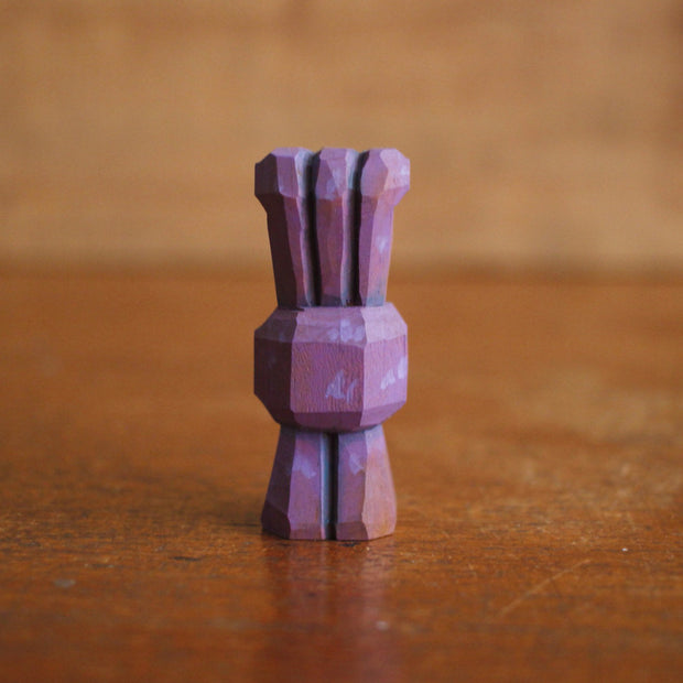 Small whittled wooden sculpture of a purple alien, with 3 eyes extended out from its head. Its body just contains its mouth, smiling with buckteeth, and it stands on 2 legs without arms.