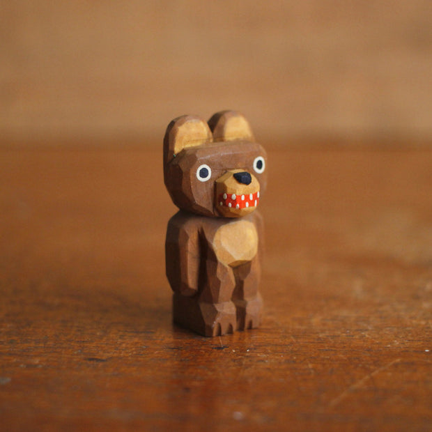 Small whittled wooden sculpture of a brown bear, standing on its hind legs like a human with its arms at its side. It has a wide red mouthed smile with small teeth.