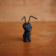 Small painted whittled wooden sculpture of a black ant with bent wires for antenna. 