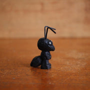 Small painted whittled wooden sculpture of a black ant with bent wires for antenna. 