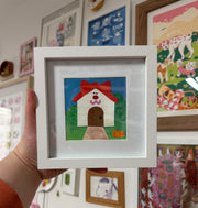 Cut paper collage of a white simplistic house with a red roof with cat ears. A cat face is on the front of the house, above a brown door. A small orange cat sits on the front lawn.