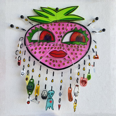 Sculpture made of assembled shrink plastic depicting a cartoon pink strawberry with large eyes and lips. Hanging from it on metal rings are tiny assorted charms.