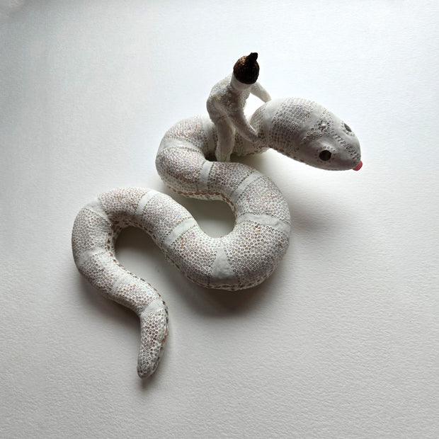 Sculpture of a white snake, seen from overhead. It has delicate patterning on its body making up its scales with thin white stripes. Riding on its neck is a woman with her arms loosely wrapped around it.