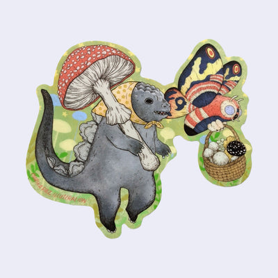 Die cut sticker of a cartoon Godzilla wearing a yellow headscarf and holding a large red mushroom over its shoulder. A Mothra flies along side while holding a basket of mushrooms.