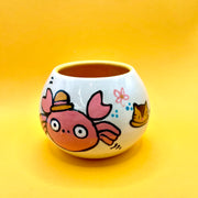 Small high walled bowl with a slightly tapered top with illustrations on it of a crab with large cartoon circular eyes and a small straw hat.