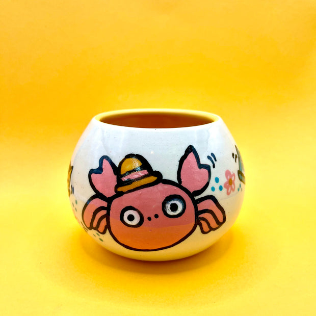 Small high walled bowl with a slightly tapered top with illustrations on it of a crab with large cartoon circular eyes and a small straw hat.