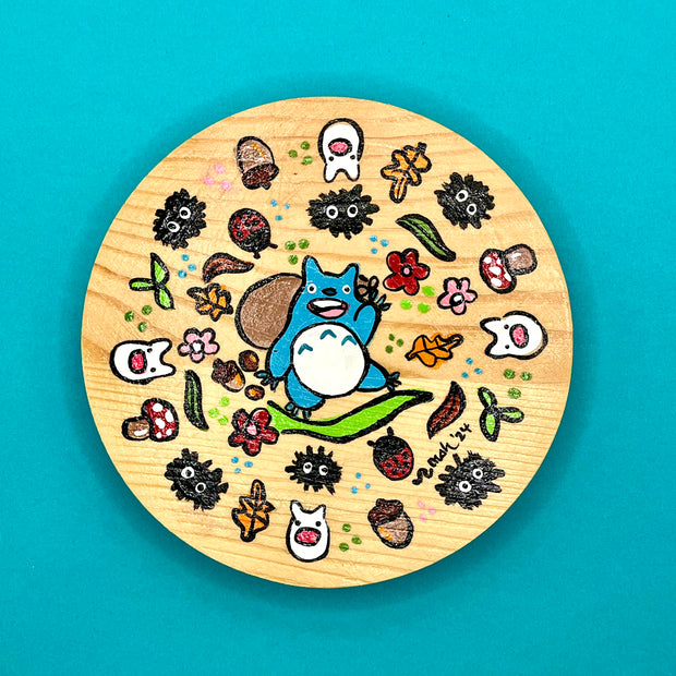 Painting on a circular wooden panel, with a stylized Blue Totoro from My Neighbor Totoro in the center. It carries a brown sack and stands atop of a leaf. Surrounding it are small bugs, autumn foliage and soot sprites.