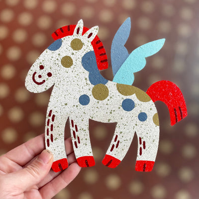 Die cut flat paper sculpture of a white horse with gold and blue spots, red mane and blue wings. It has a simplistic smiling face.