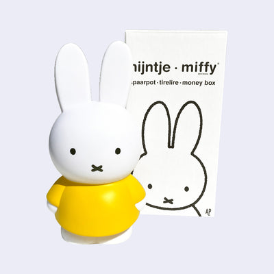 Coin bank shaped like Miffy, wearing her standard yellow dress. She stands next to the product packaging.