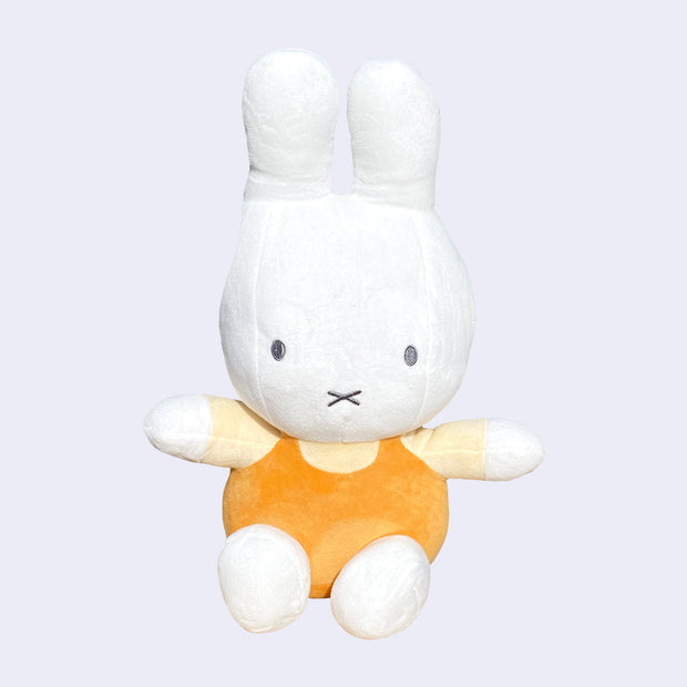 Plush doll of a white bunny with a large head and arms outspread. It wears a light. yellow shirt and orange overalls and sits.