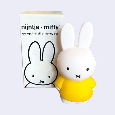 Coin bank shaped like Miffy, wearing her standard yellow dress. She stands next to the product packaging.