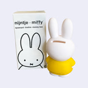 Coin bank shaped like Miffy, wearing her standard yellow dress, the back of her head has a coin slot. She stands next to the product packaging.