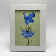 Framed painting of a blue butterfly flying over a single blue flower with many thistles coming out from the stem and around the blossom. Background is a sage green.