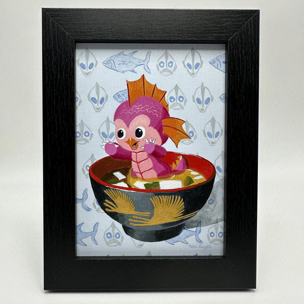 Painting of a pink mini kaiju with a cute expression and orange fins. It sits in a both of miso soup. Background is light blue with a repeating pattern of fish and Japanese robot. Piece is in a black frame.