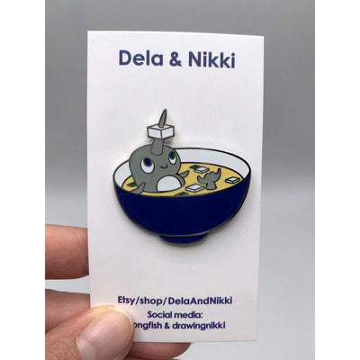 Die cut enamel pin of a narwhal sitting in a bowl of miso soup with a cube of tofu on its horn.