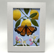 Painting of a monarch butterfly resting atop of a lemon attached to the branch with leaves and 2 more lemons behind. Below, are flower blossoms. Background is a muted purplish blue. Piece is framed.