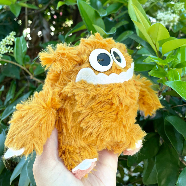 Fluffy plush doll of a yellow monster with lots of fur and a pair of big cartoon eyes and a goofy underbite smile.