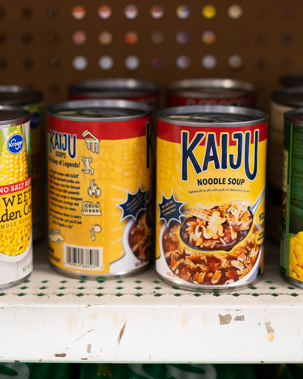 Cans on a shelf. The cans reads "Kaiju Noodle Soup" and has a photograph of a spoonful of brown soup with kaiju shaped noodles, over its bowl.