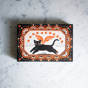 Black and white ink illustration with orange color accents of a black cat, flying over a small mountain range with wings. Border is ornate with half circles, stars and other decorative patterning.