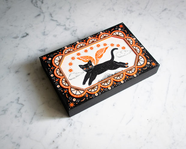 Black and white ink illustration with orange color accents of a black cat, flying over a small mountain range with wings. Border is ornate with half circles, stars and other decorative patterning.