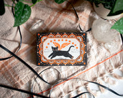 Black and white ink illustration with orange color accents of a black cat, flying over a small mountain range with wings. Border is ornate with half circles, stars and other decorative patterning.