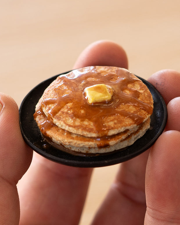 Small assembled clay sculpture of a model pancake breakfast on a plate. A pat of butter rest atop the stack with syrup poured over the pancakes. Held in the hands of the artist.