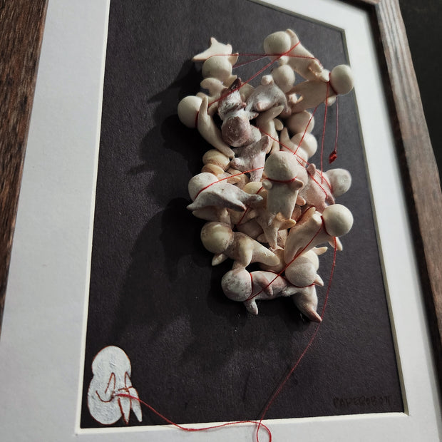 Framed sculpture of many faceless simplistic humanoid characters, all tied up with one another by a red string. The red string falls to the bottom, where a painting of a small character covering their face sits.