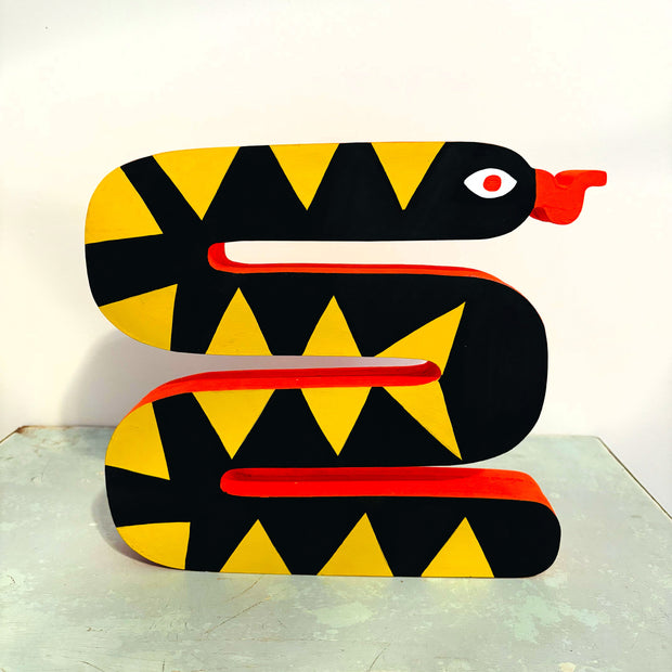 Die cut blocky wooden sculpture of a curved snake, black with yellow triangle shapes on its body. Its underbelly is a bright red and it has a wavy tongue sticking out its mouth.