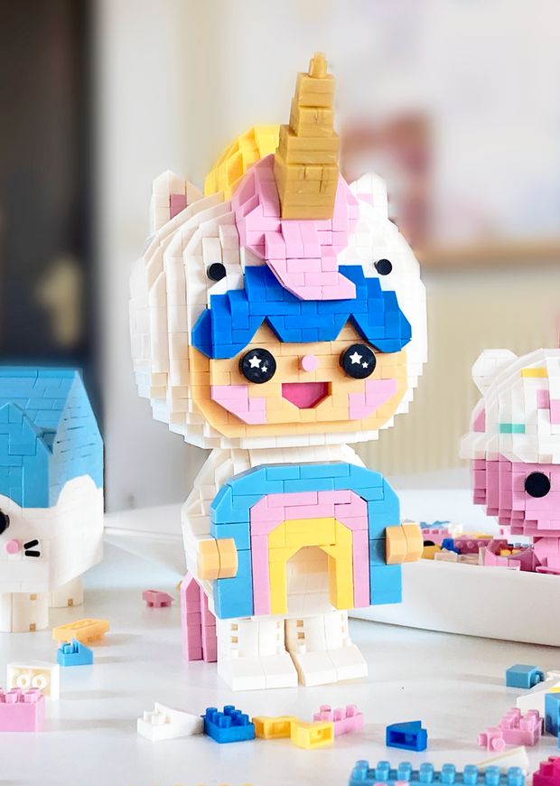 3D sculpture made out of small brick like plastic pieces of a blue haired boy wearing a white body suit and a unicorn hood hat, with pink mane and golden horn. He smiles and holds a pastel colored rainbow.