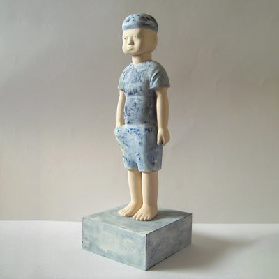 Carved wooden sculpture of a boy with a blank expression standing with one hand at his side and one reached into his pocket. He wears a t-shirt, shorts and a beanie. Colors are very muted cream and blue.