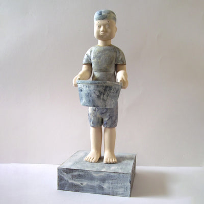 Carved wooden sculpture of a boy with a blank expression standing while holding a wide mouthed bucket. He wears a t-shirt, shorts and a beanie. Colors are very muted cream and blue.