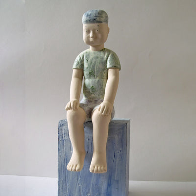 Carved wooden sculpture of a boy with a blank expression sitting on a platform with his hands on his knees and his legs dangling. He wears a t-shirt, shorts and a beanie. Colors are very muted cream, green and blue.