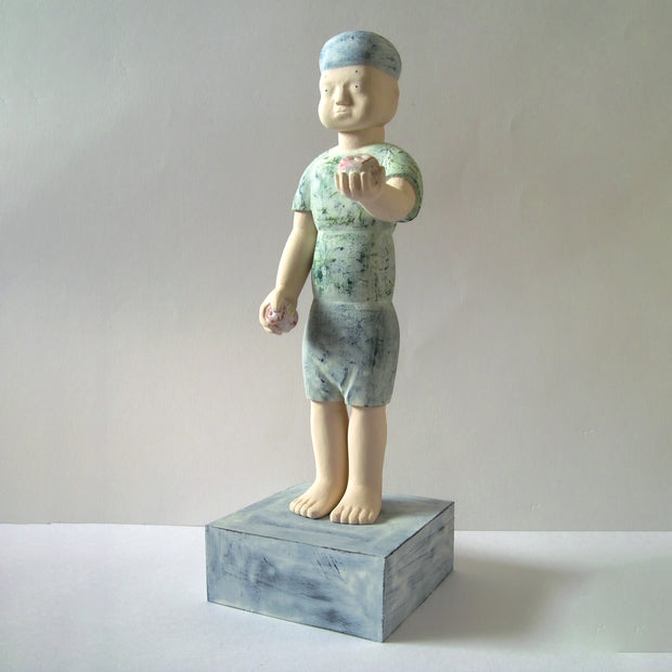 Carved wooden sculpture of a boy with a blank expression standing, holding 2 apples - one in each hand. One hand is by his side and the other is raised up, as if presenting the apple. He wears a t-shirt, shorts and a beanie. Colors are very muted cream, green and blue.