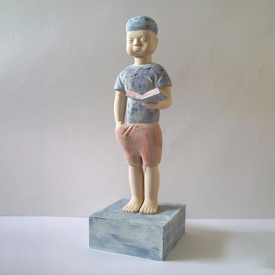 Carved wooden sculpture of a boy with a blank expression standing with one hand in his pocket and the other holding an open book. He wears a t-shirt, shorts and a beanie. Colors are very muted cream, red and blue.