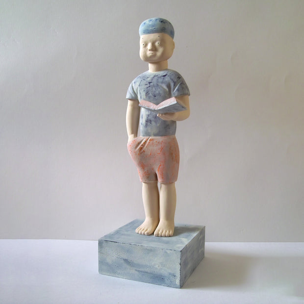 Carved wooden sculpture of a boy with a blank expression standing with one hand in his pocket and the other holding an open book. He wears a t-shirt, shorts and a beanie. Colors are very muted cream, red and blue.