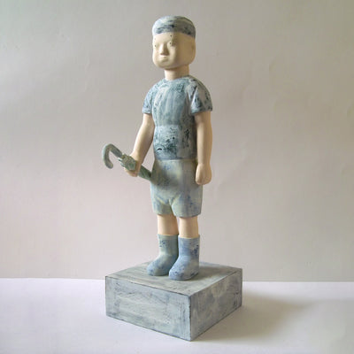 Carved wooden sculpture of a boy with a blank expression standing and holding a closed umbrella by his side. He wears a t-shirt, shorts, rainboots, and a beanie. Colors are very muted cream and blue.