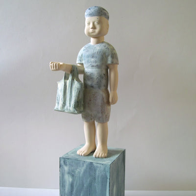 Carved wooden sculpture of a boy with a blank expression standing and holding a tote bag in the crook of his partially raised arm.. He wears a t-shirt, shorts and a beanie. Colors are very muted cream, green and blue.