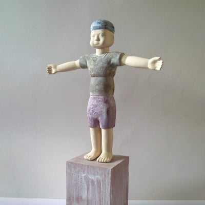 Carved wooden sculpture of a boy with a blank expression standing on a platform with both of his arms extended out as if stretching. He wears a t-shirt, shorts and a beanie. Colors are very muted cream, red, green and blue.