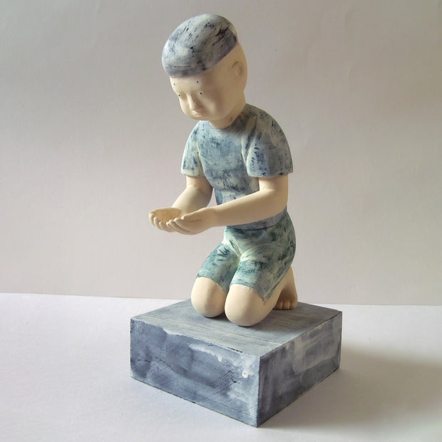 Carved wooden sculpture of a boy with a blank expression kneeling with his hands cupped in front of him. He wears a t-shirt, shorts and a beanie. Colors are very muted cream and blue.