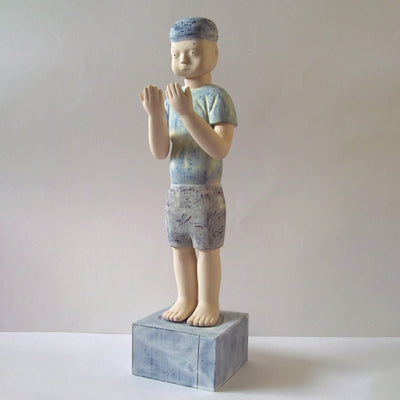 Carved wooden sculpture of a boy with a blank expression standing and looking out at his hands, which he holds up to his face. He wears a t-shirt, shorts and a beanie. Colors are very mutes cream, blues and purple.