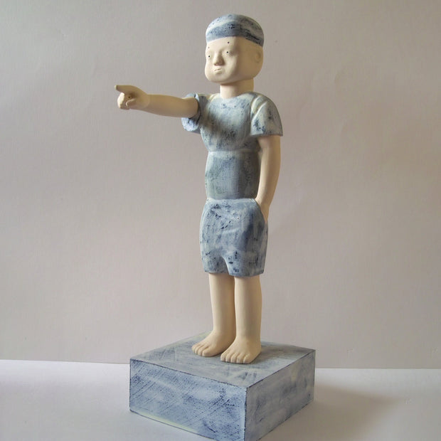 Carved wooden sculpture of a boy with a blank expression standing with one hand pointed out into the distance and the other in his pocket. He wears a t-shirt, shorts and a beanie. Colors are very muted cream and blue.