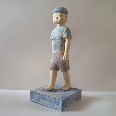 Carved wooden sculpture of a boy with a blank expression walking, with his arms at his side. He wears a t-shirt, shorts and a beanie. Colors are very mutes cream, blues and red.