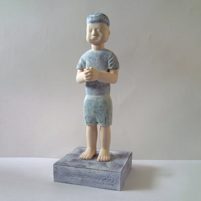 Carved wooden sculpture of a boy with a blank expression standing with his hands clasped together. He wears a t-shirt, shorts and a beanie. Colors are very mutes cream, blues and green.