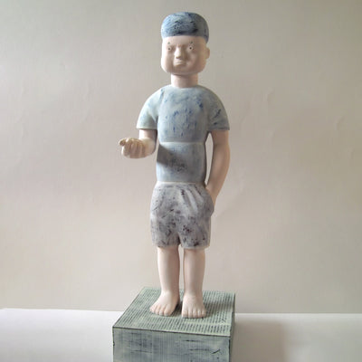 Carved wooden sculpture of a boy with a blank expression standing with one hand in his pocket and the other extended out, palm up but closed. He wears a t-shirt, shorts and a beanie. Colors are very muted cream and blue.