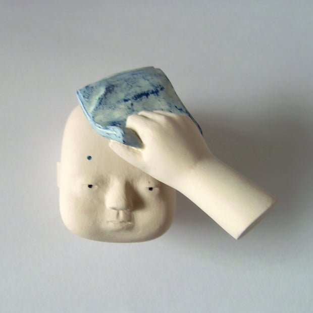 Carved wooden sculpture of a floating cream colored head with a blank expression. A floating hand holds a blue handkerchief on the head, as though soaking up any sweat.